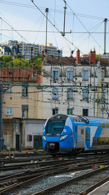 A modern double-decker train in vibrant blue and grey colors is seen approaching or passing along railway tracks in an urban setting, with overhead electrical wires visible above. Behind the train, th
