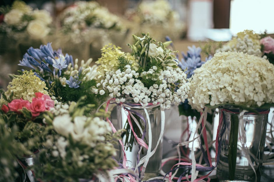 An arrangement of fresh flowers featuring white hydrangeas, white baby’s breath, and light blue irises, displayed in clear glass vases wrapped with pink and white ribbon. The flowers are arranged in