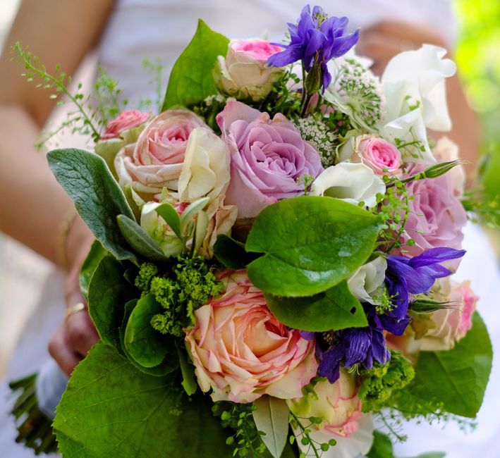 A close-up of a vibrant floral bouquet held by a person, featuring a harmonious arrangement of large, soft pink roses, creamy white roses, and delicate white lisianthus, accented with deep purple iris