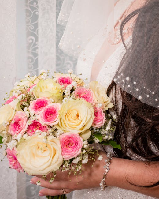 Close-up of a bride holding a floral bouquet featuring large cream and soft pink roses, complemented by smaller white filler flowers and greenery. The bouquet is wrapped with a delicate white ribbon, 