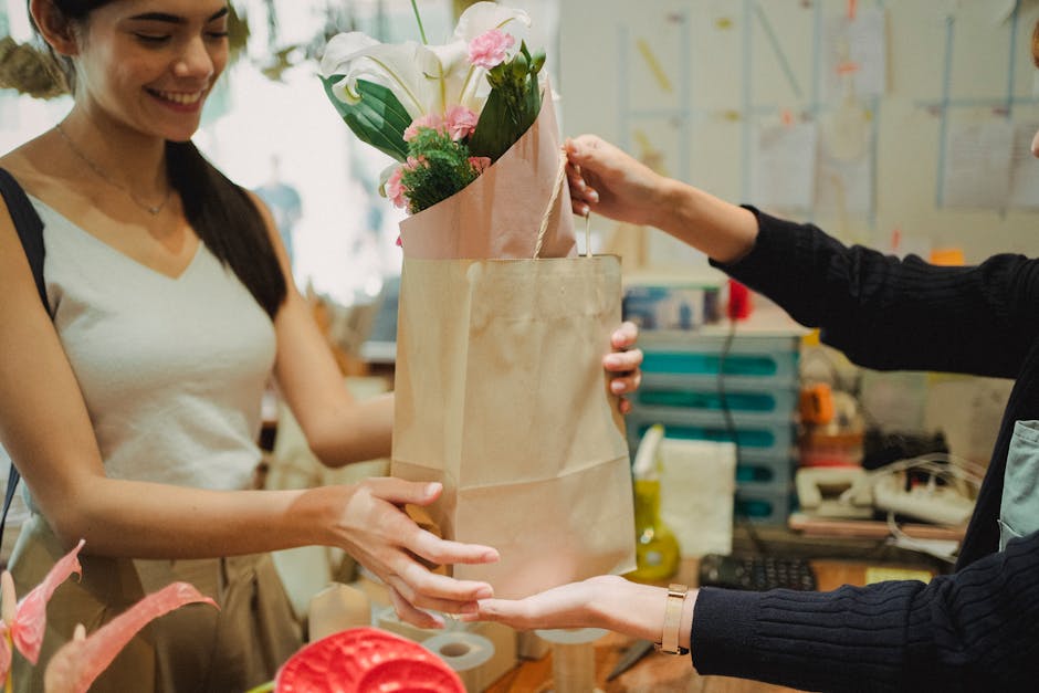 A florist at flowersbelvedere.co.uk is shown handing over a floral gift to a customer, featuring a bouquet arranged with white calla lilies, soft pink roses, and green foliage, wrapped in light pink a